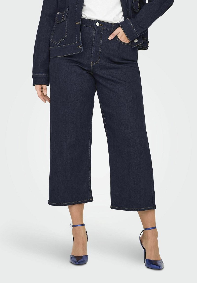 Mörkblå denimculotter med hög midja, rå kant och kontrasterande gula sömmar, i kombination med blå klackskor.