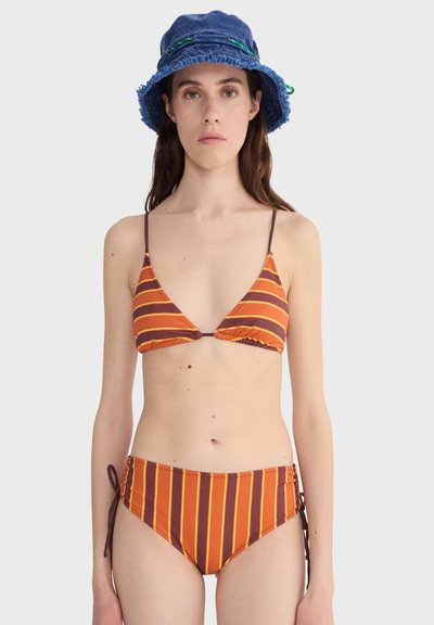 Femme portant un bikini rayé orange et marron avec des liens sur les côtés et un chapeau seau bleu, sur un fond uni clair.