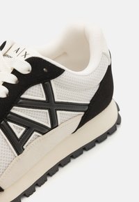 White mesh sneaker with black suede accents, featuring a raised black logo, textured detailing, and a rubber sole with traction.