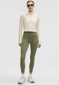 Top lung cu mânecă lungă de culoare bej deschis, cu guler scurt cu fermoar, asortat cu colanți verzi măslinii. Modelul poartă ochelari de soare și adidași albi.