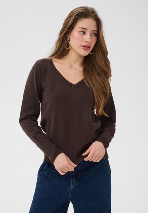Jonge vrouw met lang bruin haar draagt een donkerbruine V-hals trui, blauwe spijkerbroek, gouden oorringen en een delicate gouden ketting.