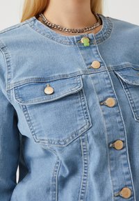 Vrouw in een lichtblauwe denimjacket met gouden knopen en gelaagde zilveren en groene kralenkettingen.