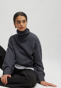 Dunkelgrauer, übergroßer Rollkragenpullover mit gerippten Bündchen. Weiche Textur, lockerer Schnitt, über einem weißen Hemd getragen. Model sitzt in lässiger Pose.