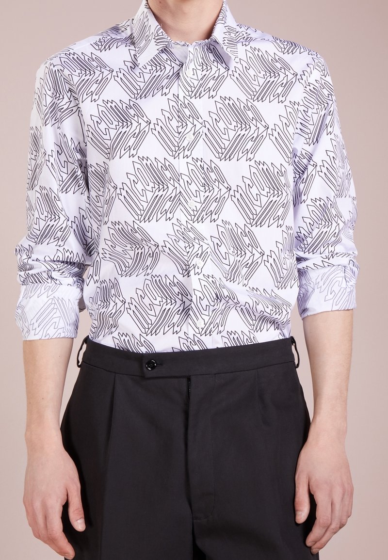 Homme portant une chemise blanche à manches longues avec des motifs géométriques abstraits noirs et un pantalon noir taille haute.