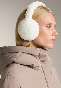 Vita konstpälsöronmuffar med en texturfinish och en vadderad pannband, bärs över en beige puffjacka i en sidoprofilvy.