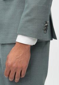 Gai pelēks uzvalka žakets ar teksturētu audumu. Piedurknei ir balts apkaklis un četri tumši pogas. Roku ir atvieglināti pie ķermeņa.