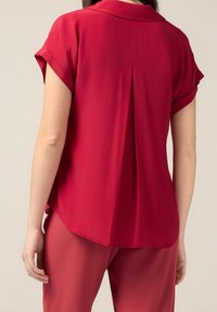 Femme portant un chemisier rouge à manches courtes amples avec des manches retroussées et un pantalon rouge assorti, vue de dos.