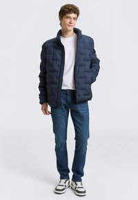 Marineblaue Steppjacke mit Reißverschluss, gesteppter Struktur und hohem Kragen. Kombiniert mit blauen Jeans und weißen Turnschuhen.