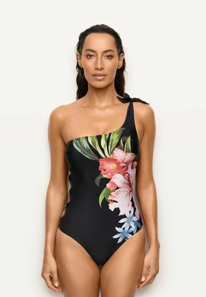 Femme portant un maillot de bain noir asymétrique avec un grand imprimé floral tropical en rose, vert et bleu, debout devant un fond clair uni.