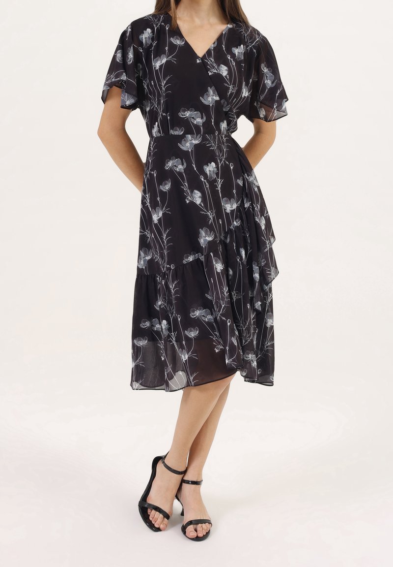 Femme portant une robe portefeuille midi noire avec des motifs floraux blancs et des manches volantes, associée à des sandales à talons noires avec des brides.