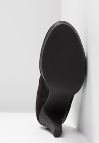 Chaussure à talon haut en daim noir avec un talon texturé gris et une semelle en caoutchouc noir lisse, positionnée sur un fond clair.