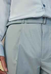 Lysblå bukser med en glat tekstur, der har en slim fit, folder og en sølvmetal spænde i taljen. En matchende lysblå skjorte er synlig.