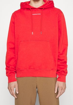 Czerwona bluza z kapturem z przednią kieszenią kangurką i napisem "SANDRO PARIS" na piersi, noszona z brązowymi spodniami.