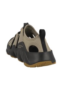 Beige Sandale mit geschlossenem Zehenbereich und offenen Seiten, schwarzer Gummisohle, strukturiertem Obermaterial, Zuglasche und kontrastierenden gelben Details.