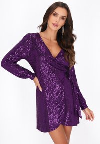 Violet sequin-mekko, jossa on pitkät hihat, V-pääntie ja kietaisumalli. Täynnä kimaltelevaa pintaa ja lyhyt helma.