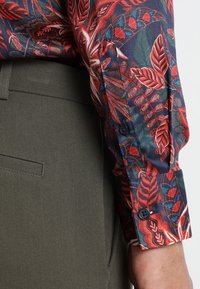 Chemise à motif floral multicolore avec des feuilles rouges, bleues et vertes. Manches longues avec poignets boutonnés, assortie à un pantalon olive.