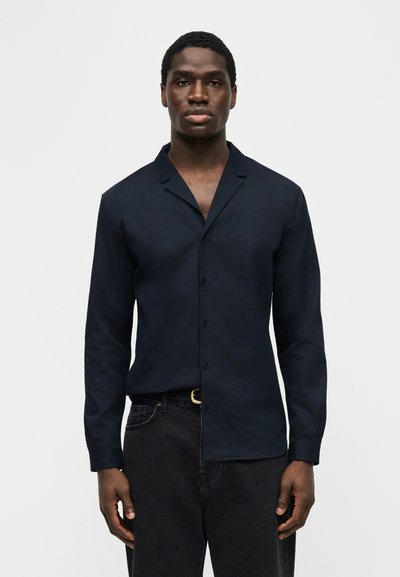 Schwarzer Mann, der ein dunkelblaues, langärmeliges Hemd mit Knöpfen und schwarze Hose trägt, steht mit entspannten Armen an den Seiten vor einem weißen Hintergrund.