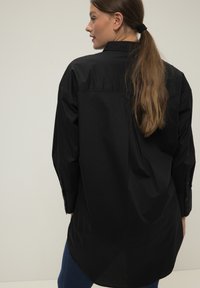 Chemise noire à manches longues avec un col, des poignets boutonnés et un ourlet légèrement arrondi. Tissu lisse avec une coupe décontractée.