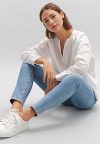 Chemise blanche à manches longues avec un col mandarin, associée à un jean slim bleu clair et des baskets blanches, assis sur un fond simple.