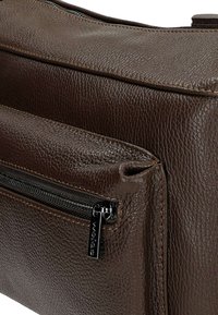 Borsa a spalla in pelle marrone con superficie tessutata, tasca frontale con zip e cuciture dettagliate lungo i bordi.