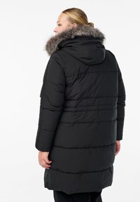 Črna puffer jakna z kapuco in umetnim krznom. Ima prešito zasnovo s horizontalnimi šivi in dolžino do kolen.