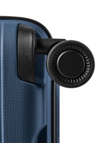 Gros plan d’un coin de valise rigide bleue avec une surface texturée et une seule roue noire rotative attachée.