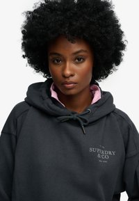 Mørkegrå hættetrøje med snøre, funnelhals og "Superdry & Co" logo. Har et pink lag synligt ved kraven. Blødt, tekstureret stof.