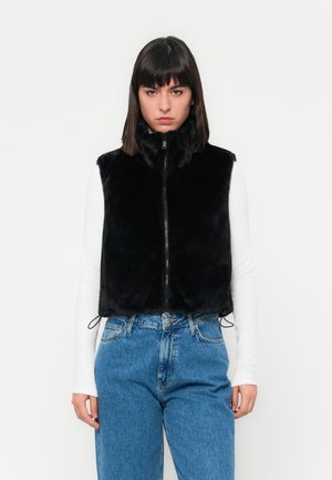 Femme se tenant face à l'avant, portant un gilet zippé en fausse fourrure noire par-dessus un haut à manches longues blanc et un jean taille haute bleu.