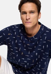 Hombre con ojos azules que lleva una camisa azul marino con un patrón de pequeños peces blancos y dorados, sentado frente a un fondo claro liso.