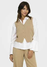 Beige vest med en tætsiddende design, fem knapper og en cropped front. Sammen med en hvid skjorte med brede manchetter og beigefarvede bukser.