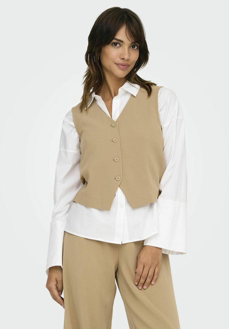 Beige vest med en tætsiddende design, fem knapper og en cropped front. Sammen med en hvid skjorte med brede manchetter og beigefarvede bukser.