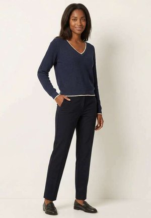 Femme debout, main dans la poche, portant un pull col en V bleu marine avec bord blanc, pantalon noir et mocassins noirs sur un fond uni.