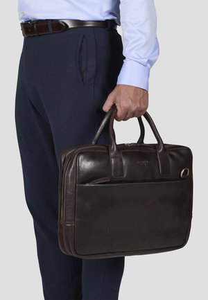 Dark brown leather briefcase with top handles, a front zip pocket, and a smooth texture. Held by a person in dress pants and shirt.