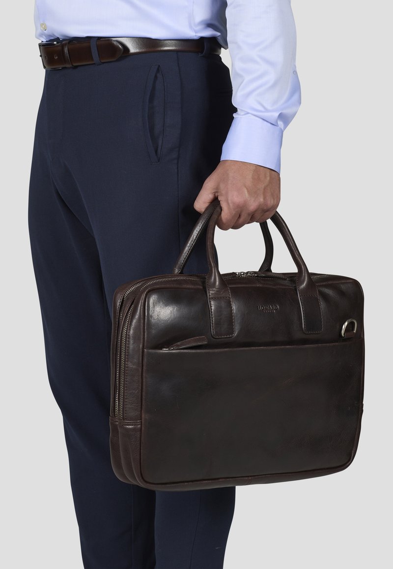 Ciemnobrązowa skórzana teczka z uchwytami u góry, przednią kieszenią na zamek oraz gładką teksturą. Trzymana przez osobę w eleganckich spodniach i koszuli.