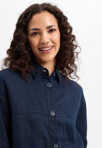 Navy blue button-up shirt in lightweight fabric, featuring a collar, chest pocket, and two buttons on the front. Soft texture and relaxed fit.