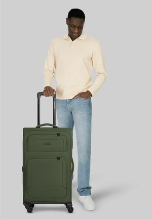 Hombre con camisa de manga larga beige y jeans sosteniendo el asa de una gran maleta verde con ruedas sobre un fondo gris.