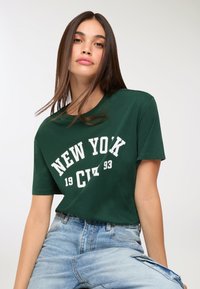 Giovane donna con lunghi capelli castani che indossa una t-shirt verde con la scritta "New York City 1993" e jeans di un blu chiaro su sfondo bianco.