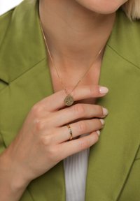 Zlatý náhrdelník s textúrovaným kruhovým príveskom, nosený spolu s jednoduchým zlatým prsteňom. Ruka spočíva na olivovo zelenom saku.