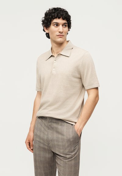 Άνδρας με κατσαρά μαλλιά, που φοράει μπεζ κοντομάνικο πόλο και γκρι καρό παντελόνια, στέκεται με το ένα χέρι στην τσέπη.