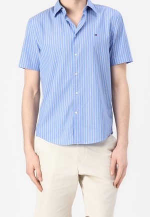 Mand iført en lyseblå kortærmet skjorte med hvide lodrette striber og beige bukser, stående foran en ensfarvet baggrund.