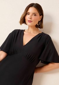 Robe noire avec un décolleté en V, des manches courtes évasées et des détails froncés à la taille, fabriquée en tissu lisse et léger.