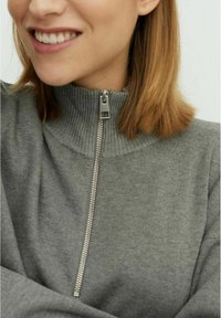 Grijze zip-up trui met een hoge geribde kraag, voorzien van een zilveren rits. Zachte textuur en getailleerd ontwerp zichtbaar in close-up opname.