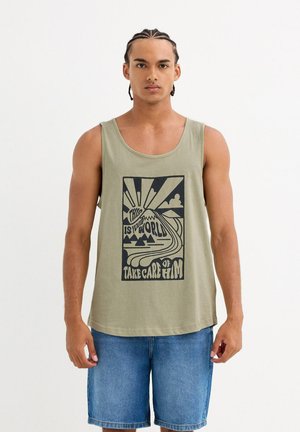 Hombre con cabello trenzado que lleva una camiseta sin mangas verde oliva con un gráfico de sol, montañas y el texto "Este es tu mundo, cuídalo", y pantalones cortos de mezclilla.
