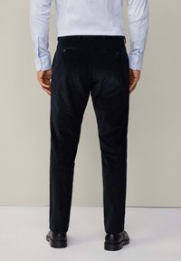 Pantaloni in corduroy neri con vestibilità slim, dotati di tasche posteriori e una texture liscia. Il modello indossa scarpe nere e una camicia azzurra chiara.