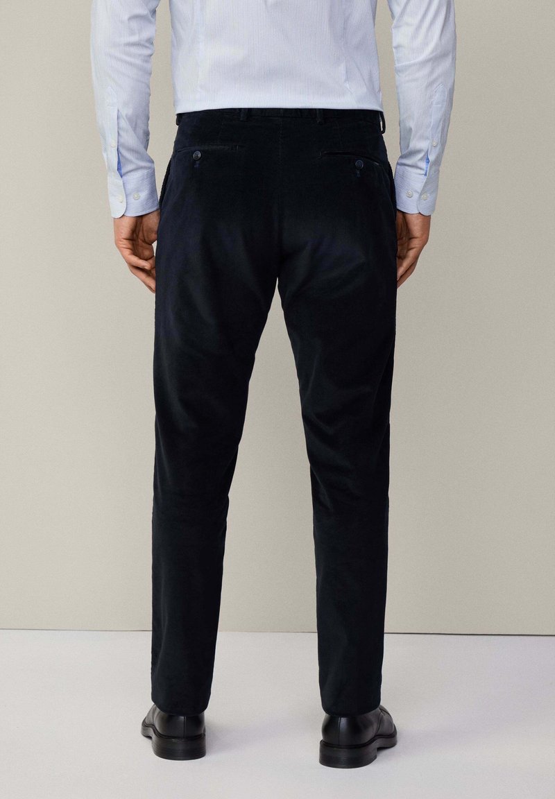 Pantalon en velours côtelé noir à coupe slim, avec des poches arrière et une texture lisse. Le modèle porte des chaussures noires et une chemise bleu clair.