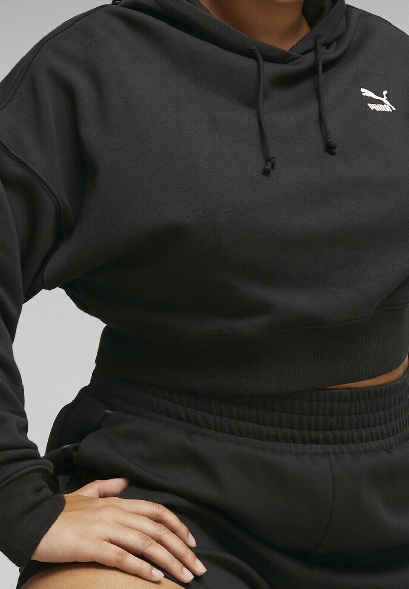 Personne portant un sweat à capuche court noir Puma et un pantalon noir assorti, la main reposant sur la cuisse sur un fond uni.