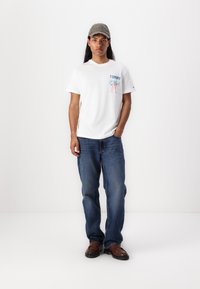 T-shirt blanc à manches courtes avec un graphique "TOMMY", texte bleu et design multicolore. Porté avec un jean bleu et des chaussures brunes. Casquette grise.