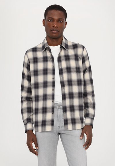 Svart och vit rutig flanellskjorta med knäppning, långa ärmar och en ficka, bärs över en vit T-shirt och grå jeans.