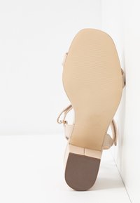 Sandale beige pour femme avec un talon bloc, une bride à la cheville et une semelle texturée, positionnée debout contre un coin blanc.