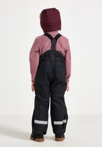 Sorte vandtætte overalls med reflekterende striber, iført over en lyserød fleece-top, suppleret af en kastanje farvet isoleret hat. Børns rygperspektiv.